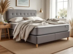 Grijze boxspring met gestoffeerd hoofdbord en beige dekbed, houten lattenbodem zichtbaar, jute vloerkleed en pampas styling