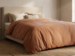 Boxspring bed met beige hoofdbord en terracotta dekbed, natuurlijke belichting in rustige slaapkamer met jute vloerkleed