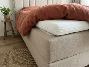 Beige boxspring bed met witte topper die van de hoek afglijdt, gestoffeerd hoofdbord en terracotta dekbed op jute vloerkleed