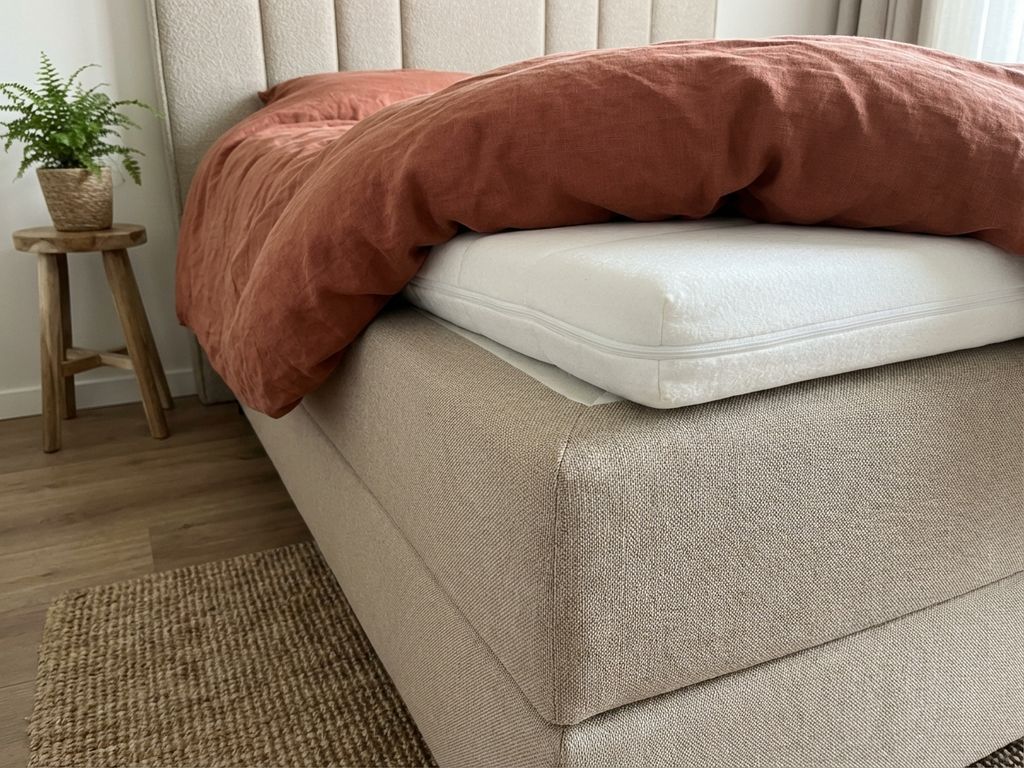 Beige boxspring bed met witte topper die van de hoek afglijdt, gestoffeerd hoofdbord en terracotta dekbed op jute vloerkleed