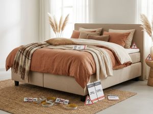 Complete boxspring bed met beige bekleding, dekbed, sierkussens en subtiele waarschuwingssymbolen voor koopadvies