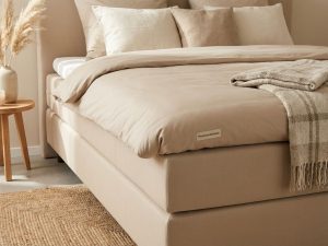 Hoogwaardige boxspring met beige dekbed zonder doorzakking, neutrale kussens en jute vloerkleed, toont goede matrasverzorging