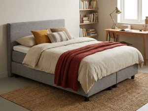 Compacte grijze boxspring met dekbed en kussens in stijlvolle studentenkamer met bureau en boekenplank