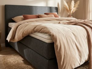 Boxspring bed met gestapelde toppers, beige dekbed, antraciet hoofdbord en terracotta sierkussens in warme slaapkamer