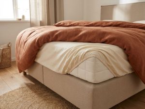 Boxspring bed met losgelaten hoeslaken in hoek, beige gestoffeerd frame, terracotta dekbed, natuurlijke verlichting